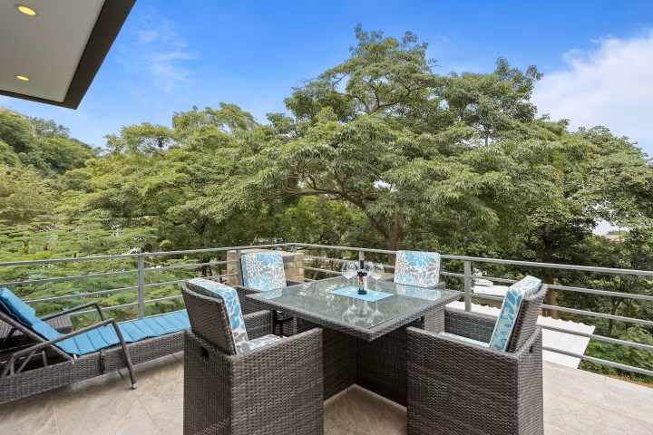 Outdoor patio with wicker chairs and a glass table on a balcony overlooking lush green trees.