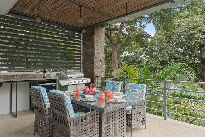 Outdoor dining area with wicker chairs, glass table, and barbecue grill under a wooden roof, surrounded by trees.