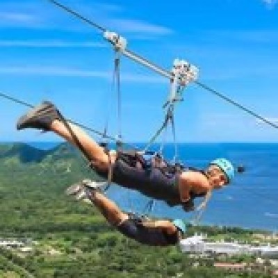 Two people zip-lining over a green landscape with ocean in the background on a sunny day.
