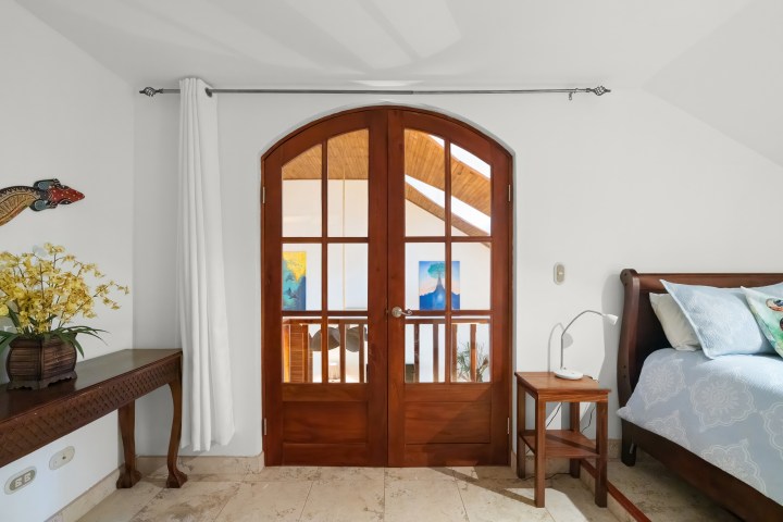 Bright room with arched wooden doors, a table with flowers, and a bed on the right.