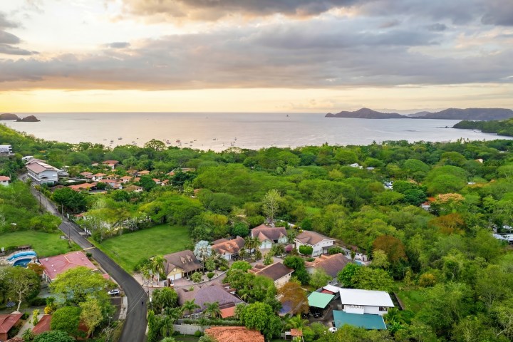 Coastal village with lush greenery and ocean view at sunset, under partly cloudy skies.