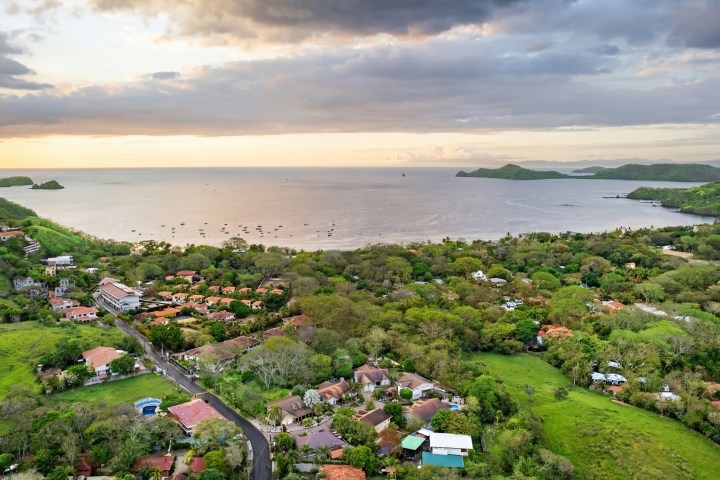 Aerial view of a coastal village with lush greenery and an ocean bay at sunset.