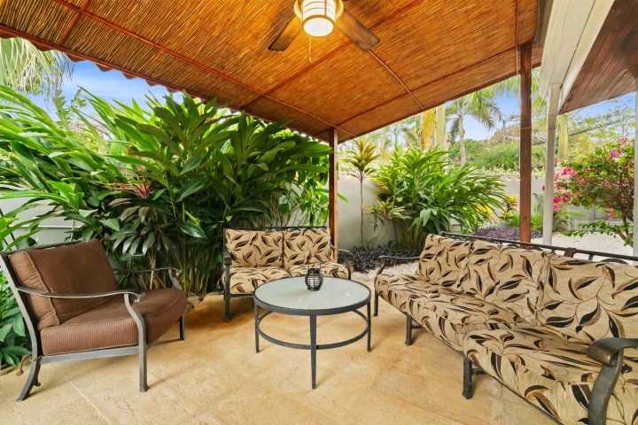 Outdoor patio with wicker furniture, cushions, and plants under a bamboo roof.