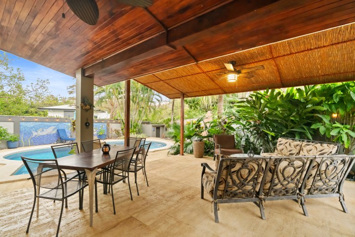 Outdoor patio with seating, dining table, ceiling fan, and view of pool surrounded by tropical plants.