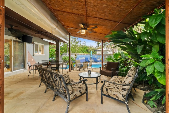 Covered patio with chairs, table, ceiling fan, and view of pool with greenery.