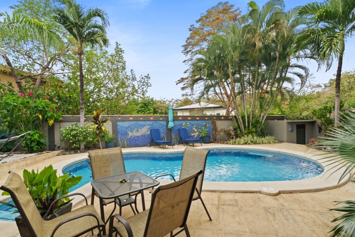 Patio with pool, palm trees, and outdoor furniture under a blue sky.