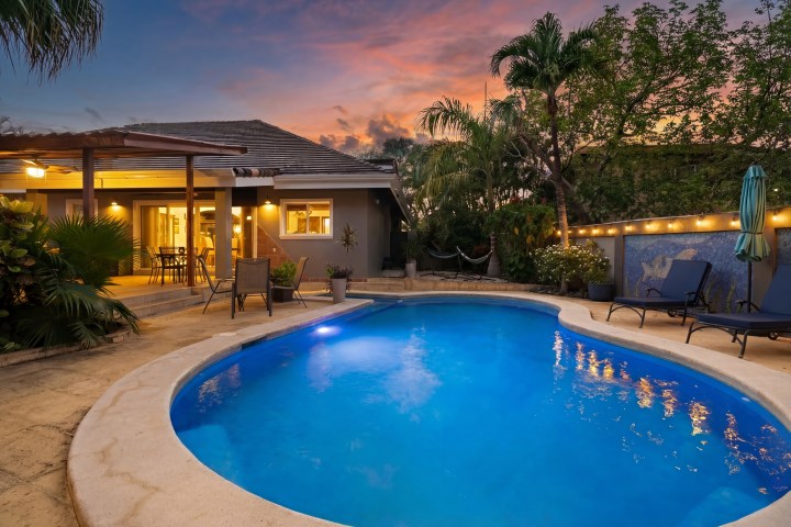 Backyard with pool, lounge chairs, and lit house at sunset.