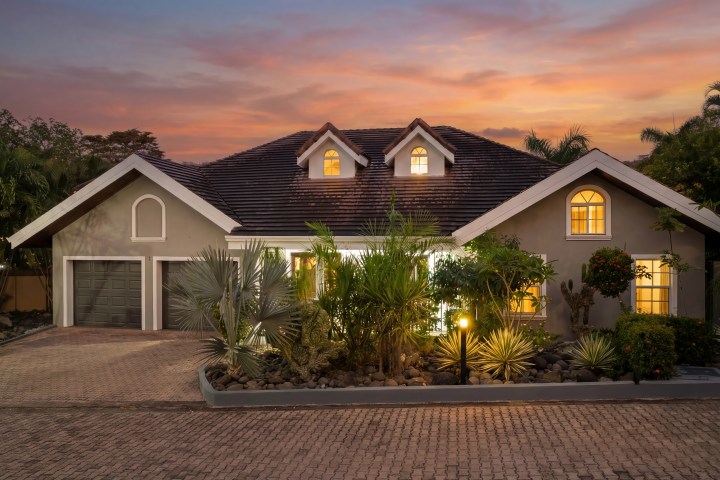 House with illuminated windows, garage, and landscaped front yard at sunset.