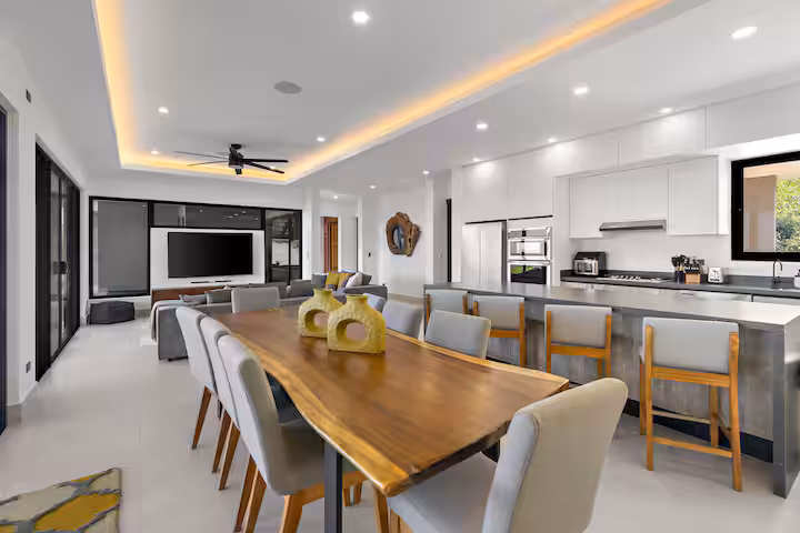 Modern open-plan kitchen and dining area with wood table, barstools, and ceiling lights.