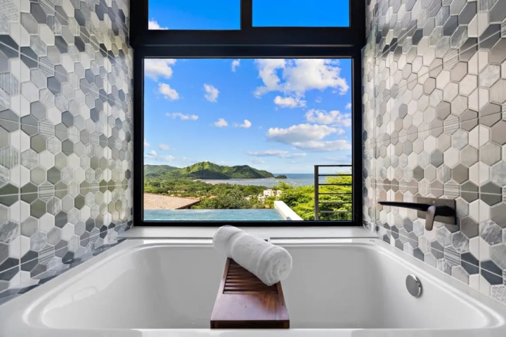 Bathtub with rolled towel, hexagonal tiles, and ocean view through a window.