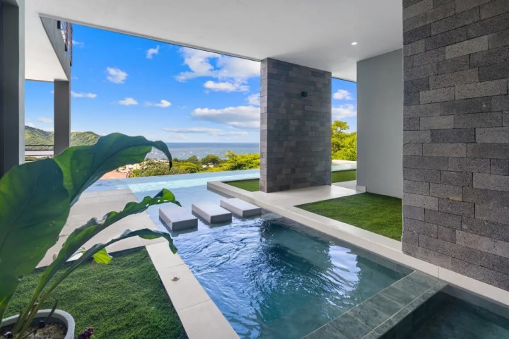 Modern patio with pool, stepping stones, and ocean view under a cloudy sky.