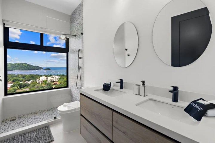 Modern bathroom with double sinks, round mirrors, and ocean view from window.