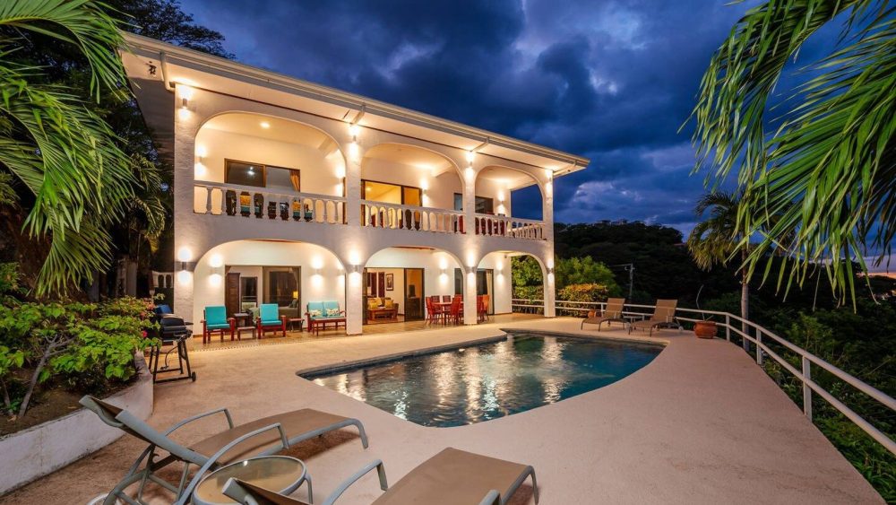 Luxury villa with pool, palm trees, and lit exterior at dusk.