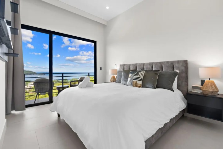 Modern bedroom with a large bed, balcony view, and ocean in the distance.