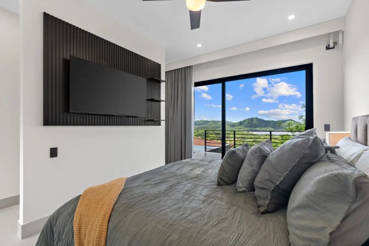 Modern bedroom with a TV, large bed, and floor-to-ceiling window overlooking a mountain view.