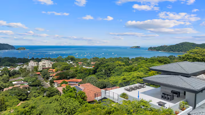 Coastal view with ocean, trees, houses, and a rooftop terrace under a blue sky.
