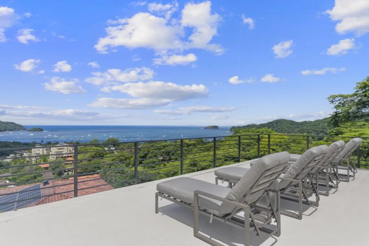 Lounge chairs on a terrace overlooking a coastal view with blue sky and clouds.