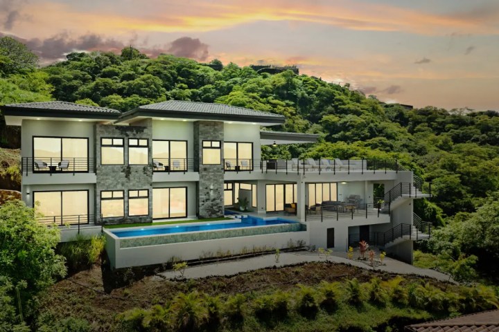 Modern hillside house with large windows, pool, surrounded by lush greenery at sunset.