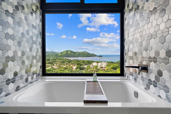 Bathtub with hexagonal tile wall and window view of ocean and hills.