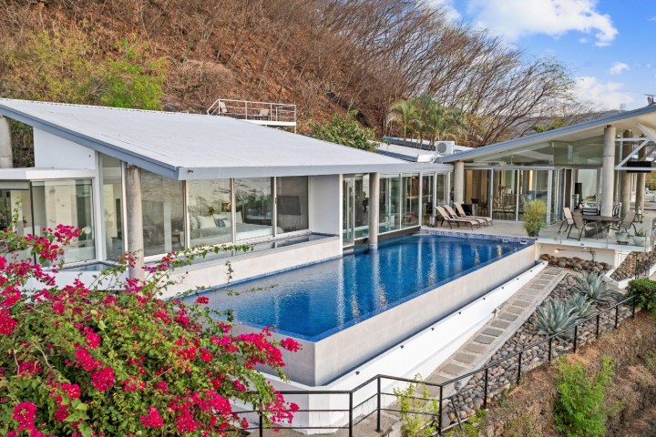 Modern house with pool, patio, and flowering plants on hillside.