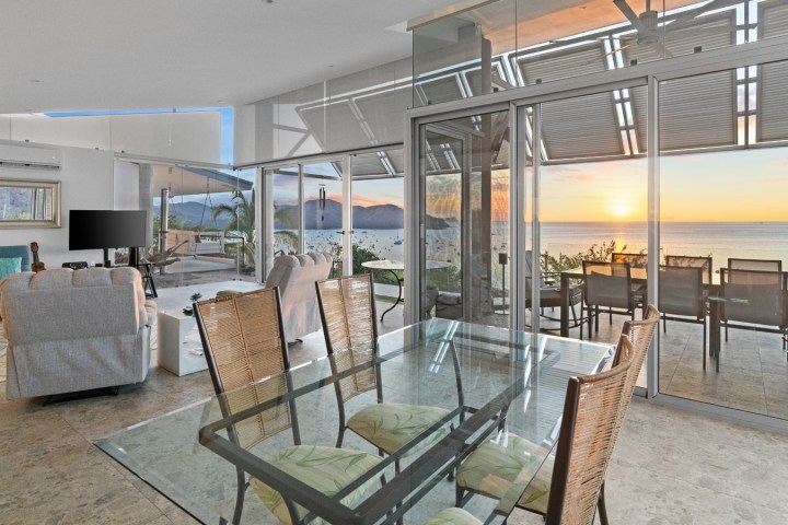 Modern living and dining area with ocean view at sunset, glass walls, and contemporary furniture.