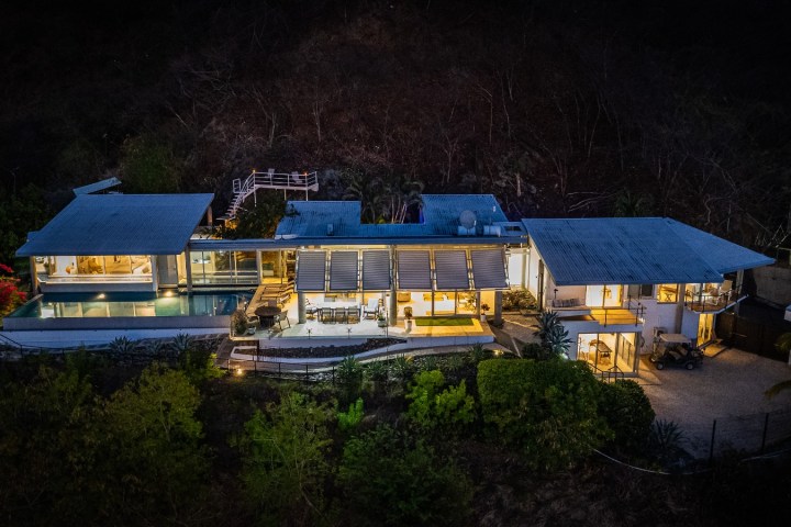 Modern house with large windows and pool, illuminated at night, surrounded by trees.