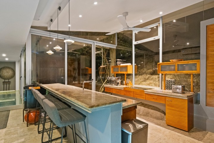 Modern kitchen with bar stools, marble countertop, and glass wall overlooking a pool.