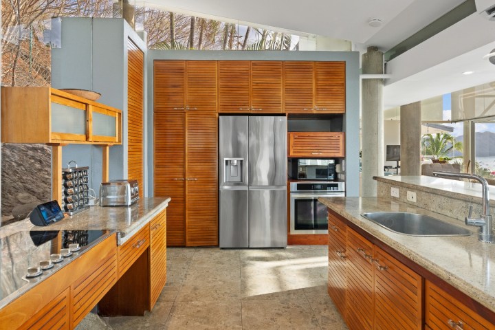 Modern kitchen with wooden cabinets, stainless steel appliances, and stone countertops, surrounded by large windows.