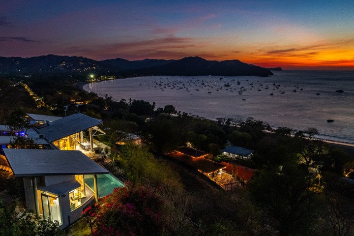 Coastal hillside at sunset with houses, ocean, and distant mountains.