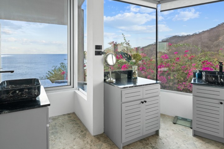 Modern bathroom with ocean view, two sinks, and flowers outside windows.
