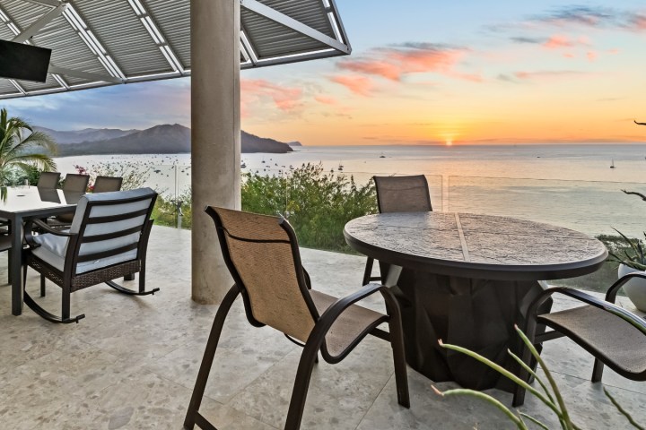 Seaside patio with tables, chairs, and ocean sunset view.