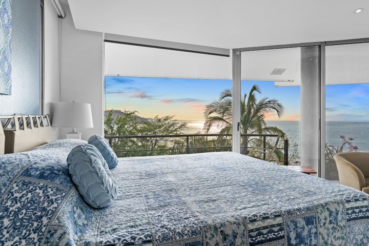 Bedroom with blue patterned bedding, large windows, ocean view, and sunset.