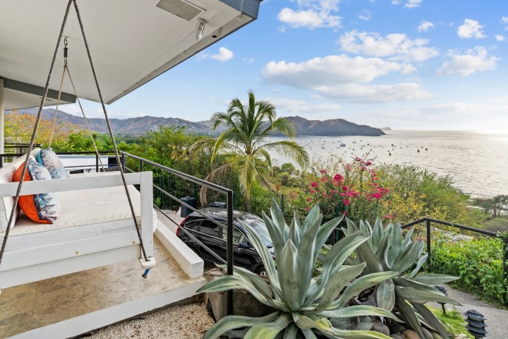 Patio with a swing seat overlooking ocean and mountains, surrounded by lush greenery.