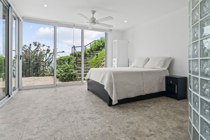 Modern bedroom with large windows, tiled floor, and plants outside.