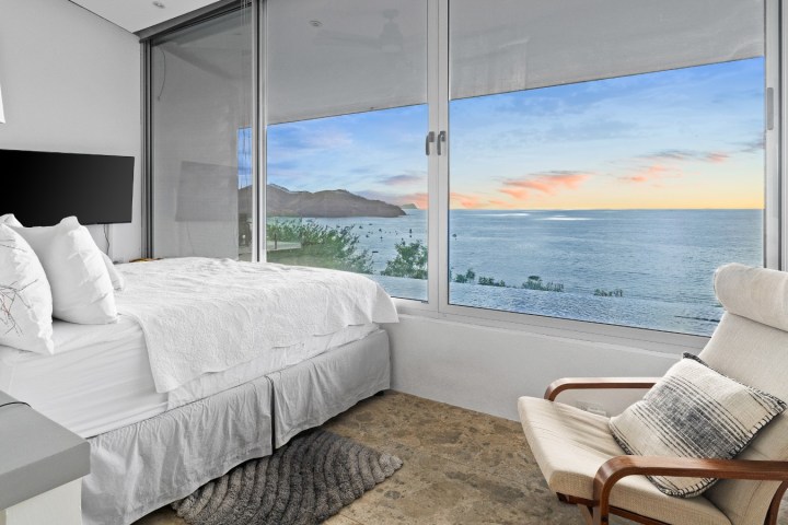 Modern bedroom with ocean view through large windows, featuring a bed and armchair.
