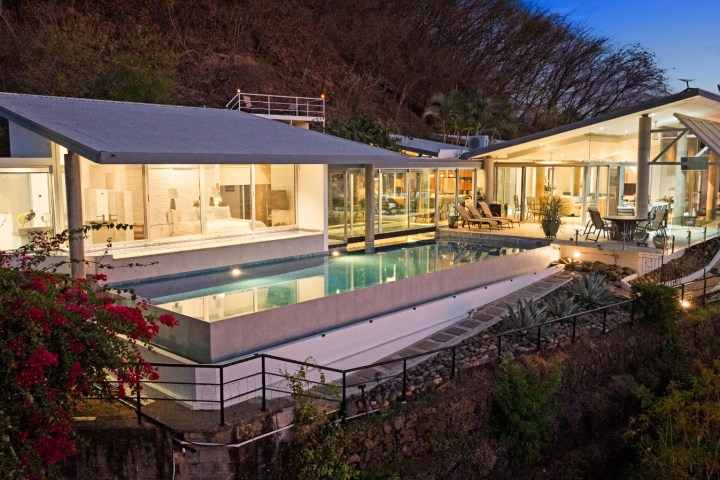 Modern hillside house with large windows, infinity pool, and deck at dusk.
