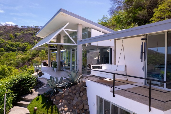 Modern hillside house with large windows and balcony, surrounded by lush greenery.