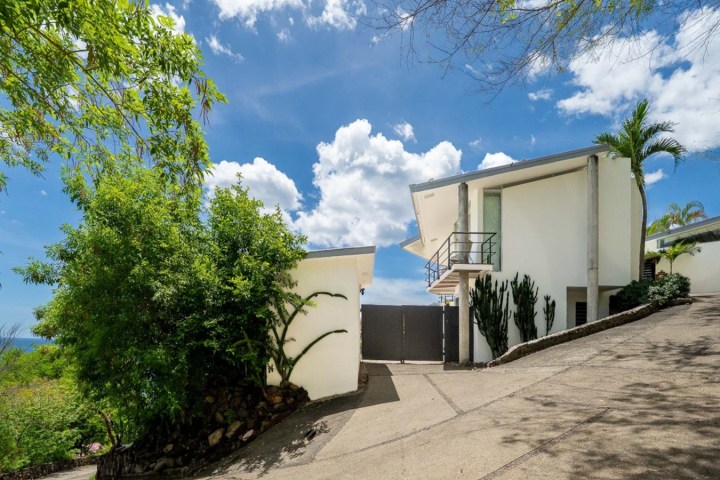 Modern white house with lush greenery and a sloped driveway under a blue sky with clouds.