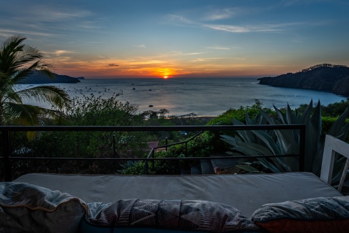 Sunset view over the ocean from a patio with a couch and palm tree.