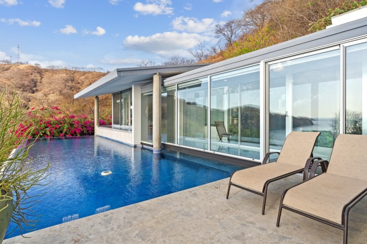 Infinity pool by a modern house with lounge chairs and ocean view.