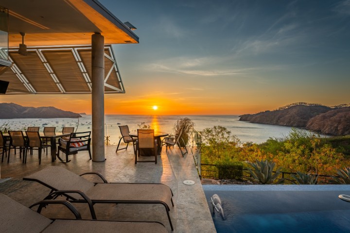 Sunset view from a patio with lounge chairs and a pool overlooking the ocean.