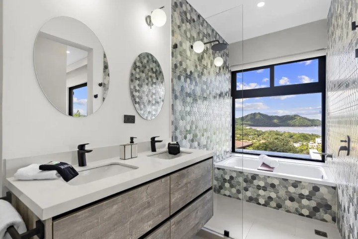 Modern bathroom with round mirrors, dual sinks, hexagonal tile accent wall, and scenic view from window.