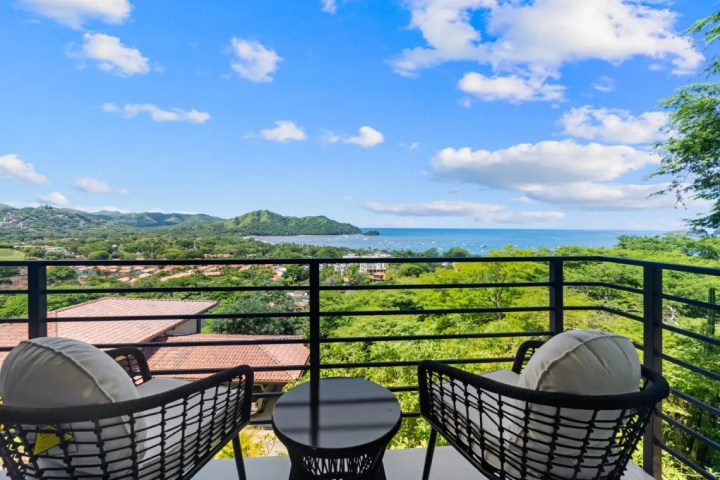 Two chairs and a table on a balcony overlooking the ocean and hills under a blue sky.