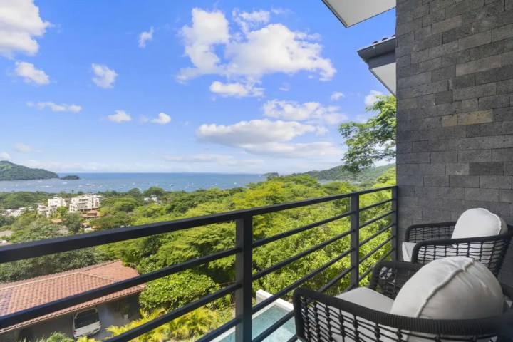Balcony view of ocean, trees, and sky with clouds.