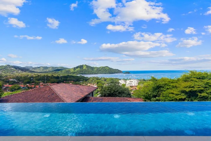 Infinity pool overlooking ocean, green hills, and blue sky with clouds.