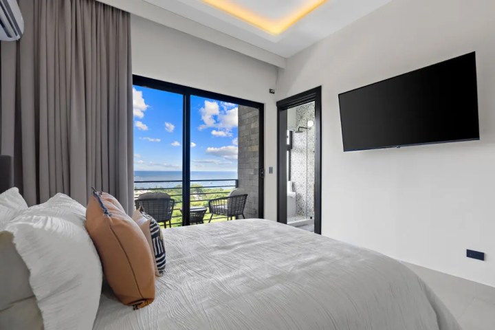 Modern bedroom with balcony view of ocean, wall-mounted TV, and neutral decor.