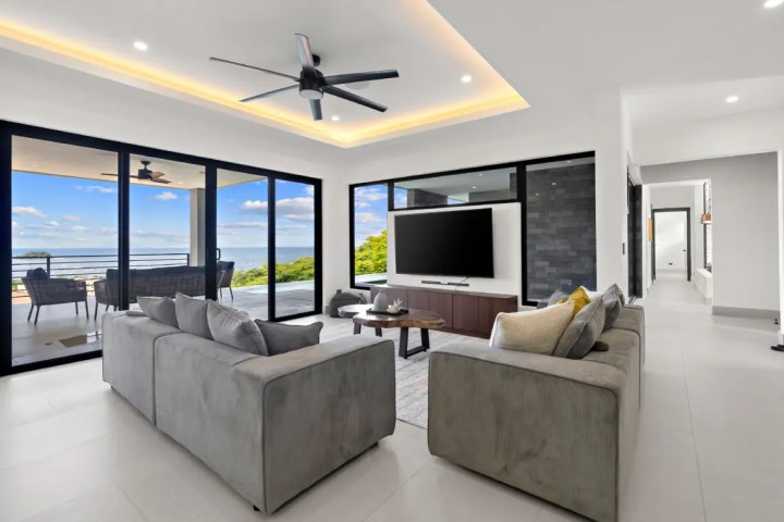 Modern living room with gray sofas, TV, large windows, ocean view, and ceiling fan.