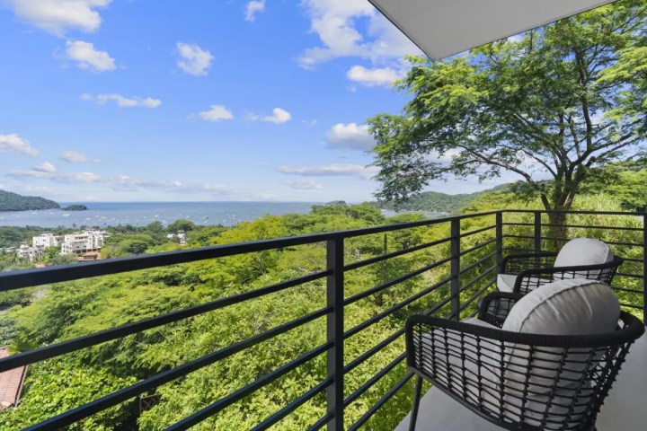 Balcony with chairs overlooking trees and ocean under a blue sky.