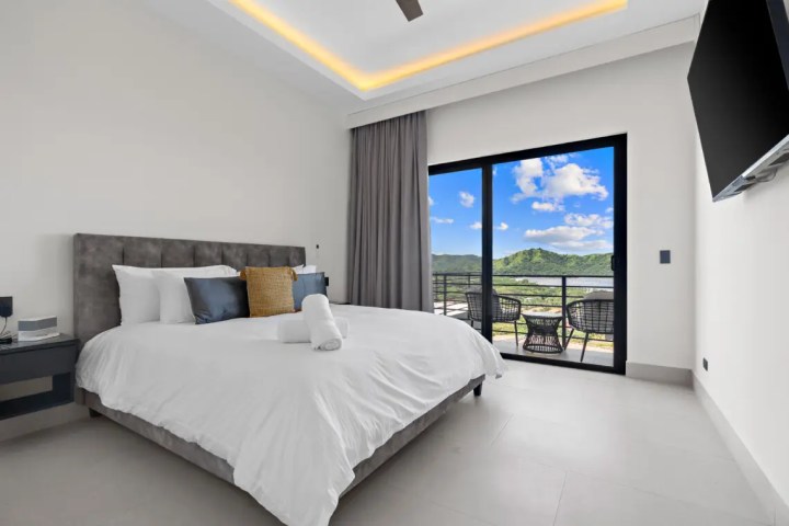 Modern bedroom with large bed, balcony view of hills, wall TV.