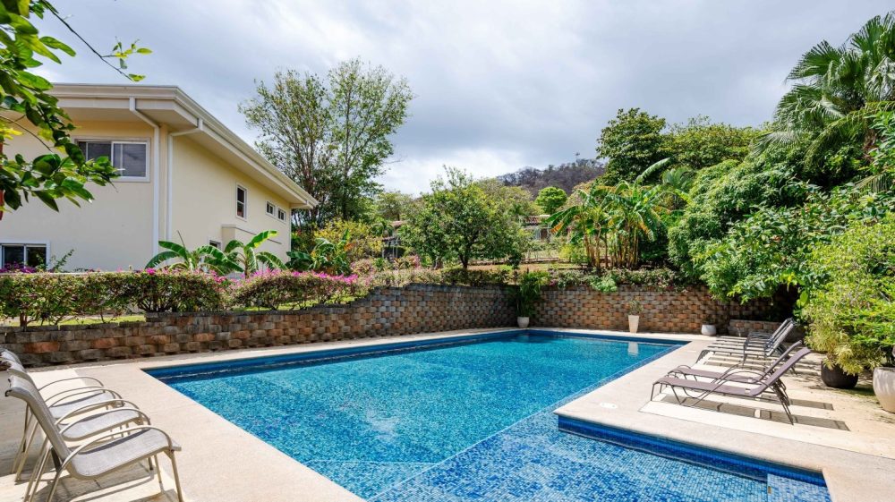 A backyard swimming pool surrounded by chairs and lush greenery.